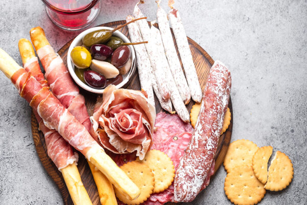 Variety of cold meat cuts and appetizers, red wine, prosciutto, jamon, salami slices, sausage, grissini, olives. Assorted mix of meat on rustic wooden board, grey stone background, top view, close-up